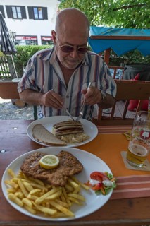 Senior citizen with sunglasses eating bratwurst in a beer garden, in front a Wiener Schnitzel with