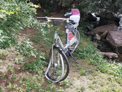 A vandalised bicycle, e-bike from a bike rental company stands in the countryside next to a river,
