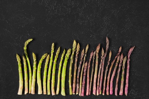 Different varieties of fresh asparagus are arranged neatly on a dark textured surface. The vibrant
