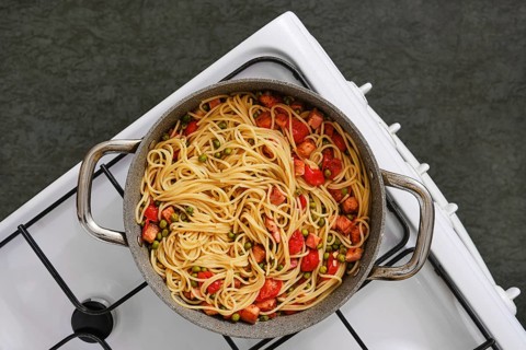 Overhead view of a pot filled with freshly cooked pasta with bacon and tomatoes on a stovetop. The