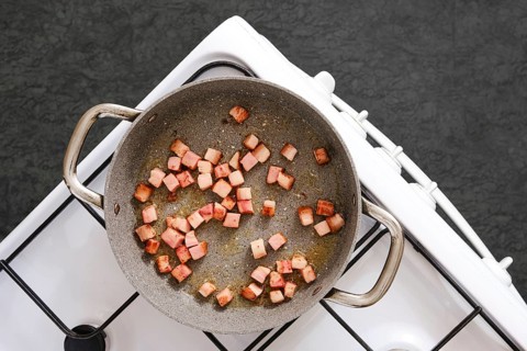 Diced smoked bacon is sizzling in a pan on a stove, browning evenly in oil. The dark textured