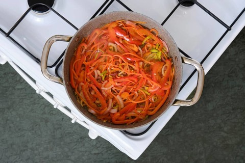 Freshly sliced bell peppers, carrot and onions simmer in a pot on the stove in a cozy kitchen