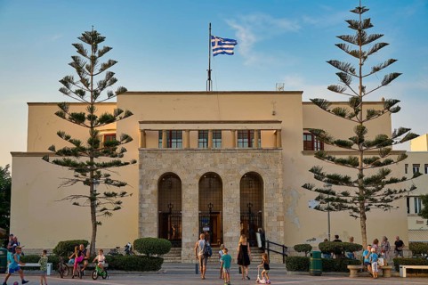 Large symmetrical building with Greek flag surrounded by people in the evening light, Kos Town,