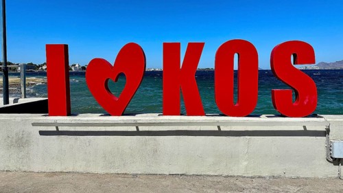 Red lettering 'I??KOS' in front of a blue sea, modern typography style under a clear summer sky,