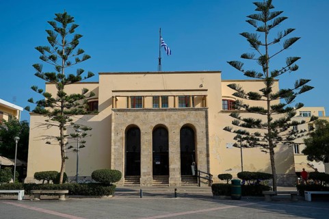 Archaeological Museum, Symmetrical beige building with Greek flag and tall trees, under clear sky