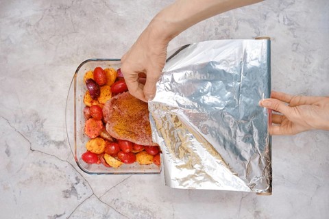 A hand gently covers a baking dish filled with seasoned turkey breast and juicy vegetables with