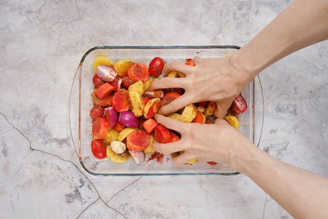 Hands mix together seasoned potatoes, tomatoes, and onions in a glass dish for a turkey breast