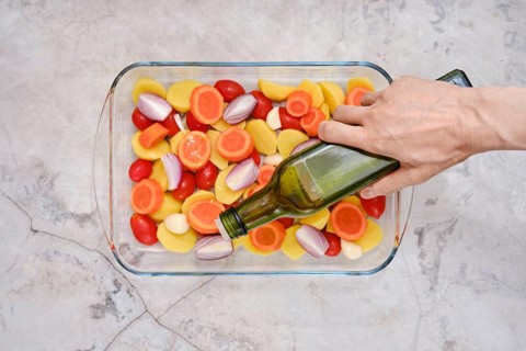 Pouring olive oil on slices of fresh vegetables including tomatoes, carrots, potatoes and onions