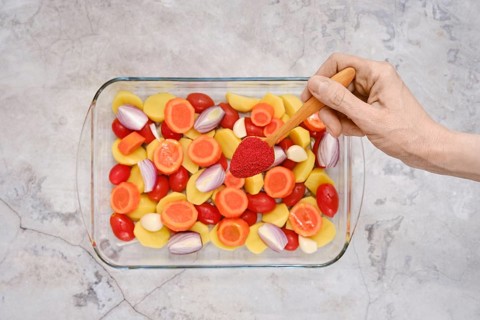 A hand holds a wooden spoon with smoked paprika above a glass dish filled with vibrant vegetables