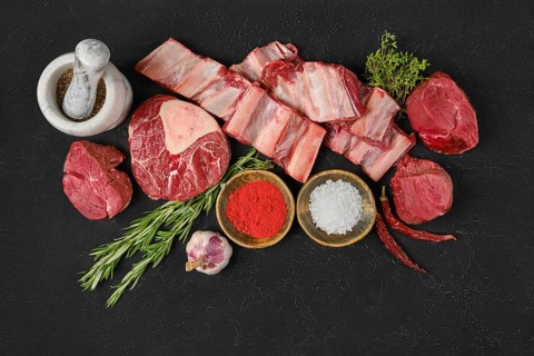 A variety of fresh cuts of beef are arranged on a dark surface, surrounded by herbs, spices, and