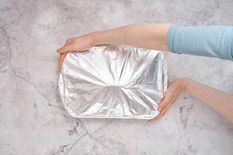 A hand holds a baking dish covered with aluminum foil, ready for the oven. The setting features a