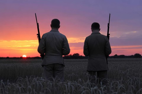 Back view of two soldiers standing in grain field watching sunset. Captures moment of reflection