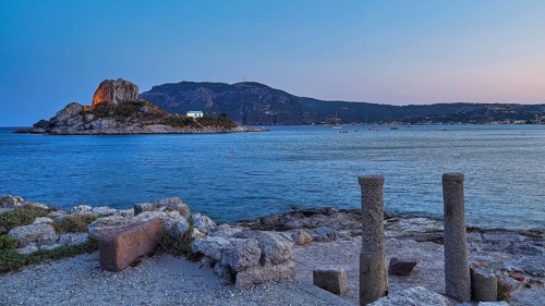 Ruins in the foreground and a calm sea with a rocky island at dusk, Agios Stefanos Beach, Agios