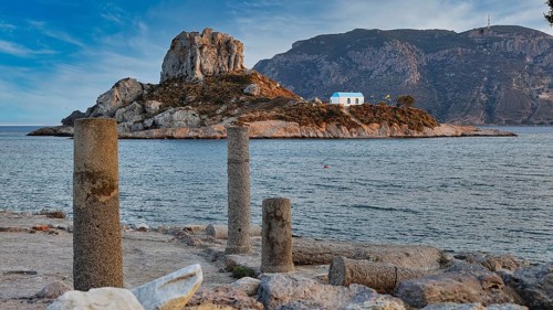 Ancient columns overlooking a rocky island with a blue house in the sea, Agios Stefanos Beach,