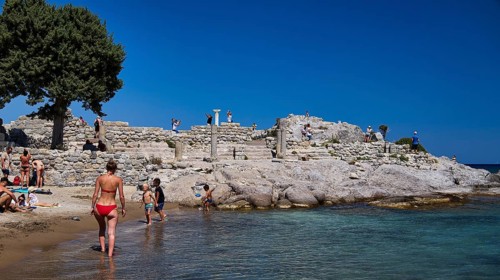 People exploring ancient ruins on the beach under a clear blue sky, Agios Stefanos Beach, Agios