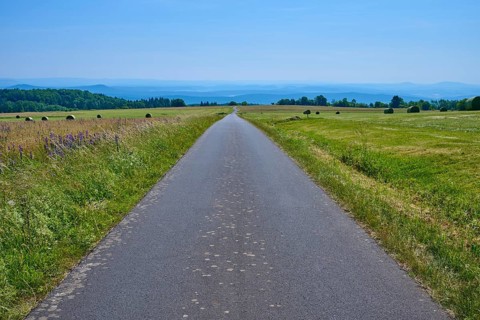 Straight road through a vast landscape of green meadows and distant hills under a blue sky,