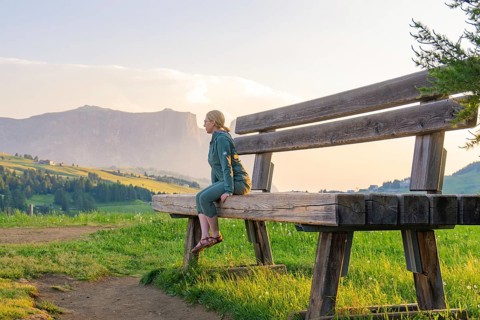 A woman sits on an oversized bench and gazes at the picturesque mountain landscape, Alpe di Siusi,