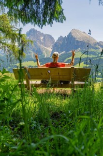 Person on a bench showing a peace sign in front of a mountain landscape, Alpe di Siusi, Dolomites,