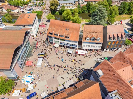A lively, summery aerial view of a square with people, market stalls and surrounding buildings, 950