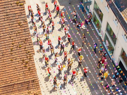Parade with people in colourful outfits on a street decorated with flags, taken from a bird's eye