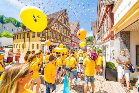 People in yellow T-shirts celebrate in a narrow street with half-timbered houses under colourful