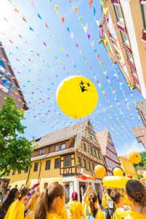 Yellow balloons rise into the sky above a busy street with historical flair and colourful pennants,