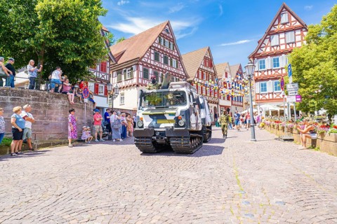 A military vehicle drives on a festive street with half-timbered buildings and people passing by,