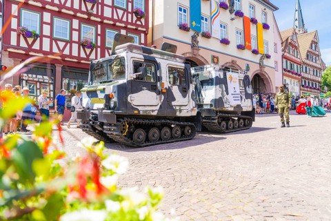 Two military vehicles in front of a row of historic half-timbered houses and surrounded by blooming