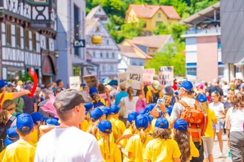 Children in yellow shirts and blue caps march with signs through a busy half-timbered street, 950