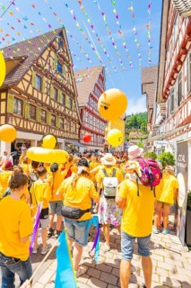 A colourful parade with yellow balloons moves through lively streets lined with half-timbered