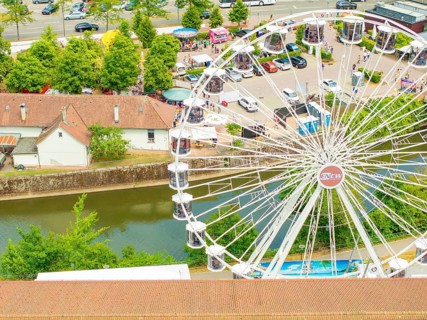 Ferris wheel stands next to a river and car parks, surrounded by trees in summer, 950 years of