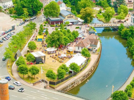 Aerial view of a small park with people, surrounded by a river and city infrastructure, 950 years