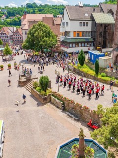 A parade moves through a market square, surrounded by half-timbered houses and many spectators in