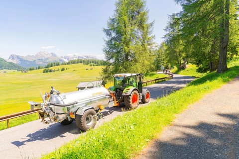 A tractor drives on a road in the middle of a green and sunny mountain landscape, Alpe di Siusi,