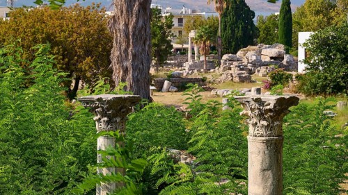 Two ornate ancient columns rise out of lush green foliage in a landscape of ruins, Ancient Agora,