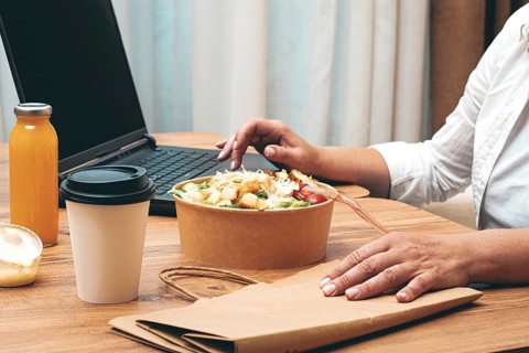 A woman is working on a laptop at home, there is food on the table, salad, coffee and juice,