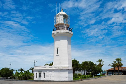 White lighthouse against a blue sky with some clouds surrounded by palm trees and vegetation,