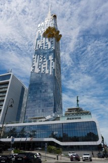A large high-rise building with a glass façade stands in an urban environment. The sky is cloudy,