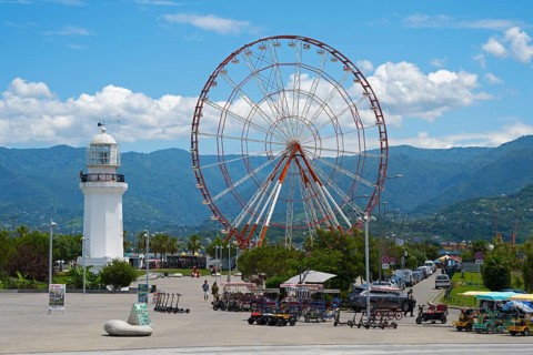 Big Ferris wheel and white lighthouse with mountains in the background under blue sky, amusement