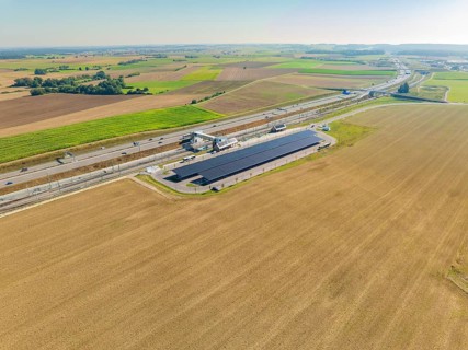 Solar system and motorway run parallel through wide fields under clear skies, e-charging park,