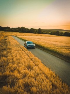 A car drives along a road surrounded by vast fields of corn with a colorful sunset in the