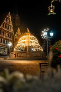 Illuminated fountain in front of half-timbered houses surrounded by festive Christmas lights at