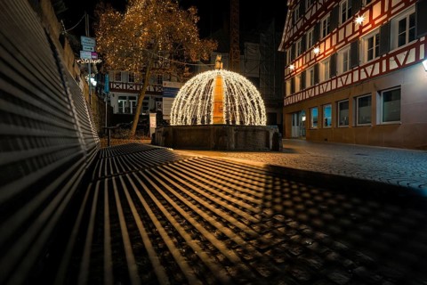 Night view of a festively illuminated fountain with shadows cast in a historic street, Calw, Black