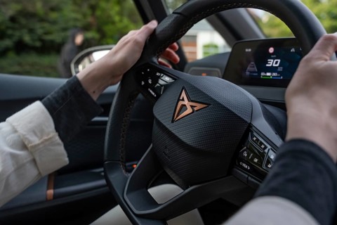A person holds a Kupra steering wheel while the driver sits in the car and shows the dashboard with