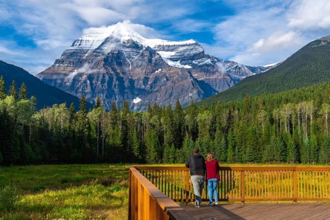Two tourists standing on a wooden deck are enjoying the breathtaking view of mount robson, the