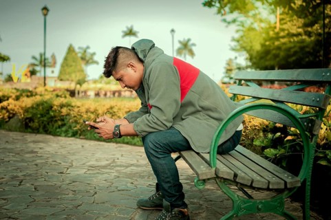 Young man in sweater sitting on a bench texting with phone. Handsome guy sitting on a park bench