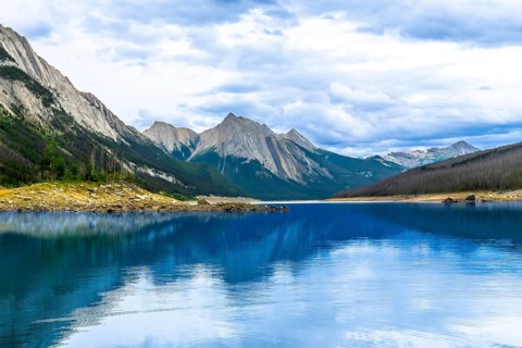 Calm turquoise water of medicine lake reflecting surrounding mountains under a cloudy sky in jasper