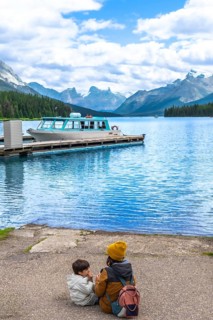 Mother and son sitting on maligne lake's shore, admiring the turquoise water and a tour boat at the