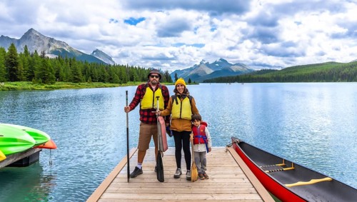 Family wearing life jackets and holding paddles, standing on a wooden dock with canoes and kayaks,