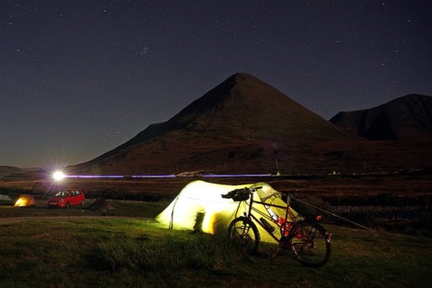 An illuminated tent next to a bicycle in front of a mountain on a clear starry night, Isle of Skye,